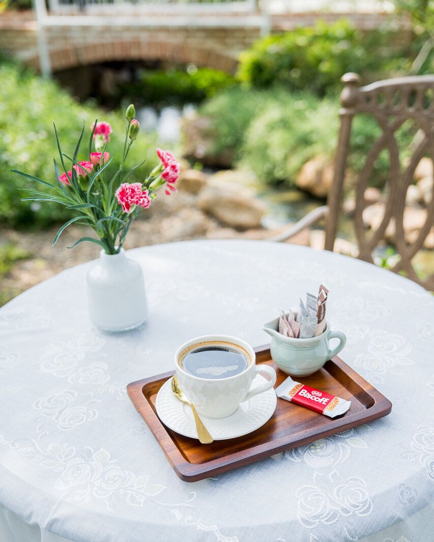 A coffee tray on a lace cloth at a garden table, with the WIROON garden behind